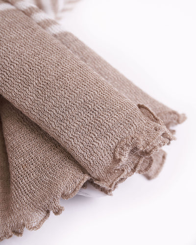 Soft textured linen scarf arranged in a loose knot, featuring a delicate beige and white striped pattern with subtle textural details. The ends of the scarf have a frayed, fuzzy finish, adding a casual yet elegant touch. The scarf is displayed against a white background, highlighting its cozy and inviting appearance